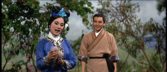 Margaret Tu Chuan in a blue top with gold embroidery sings while Kiu Chong, wearing a brown robe, looks on.
