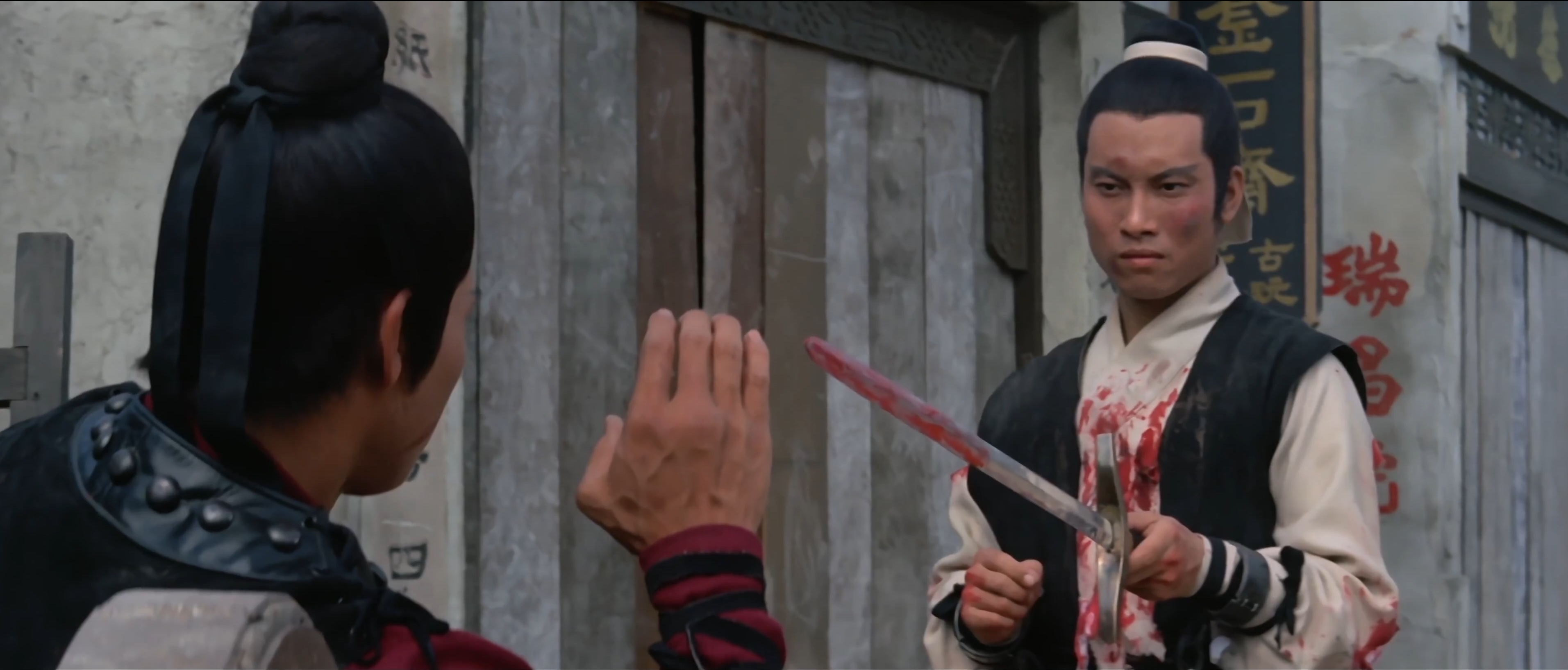 A bruised and bloody Elliot Ngok Wah holds a blood-stained sword, pointing it at Lo Lieh, who is holding his hands up in surrender.